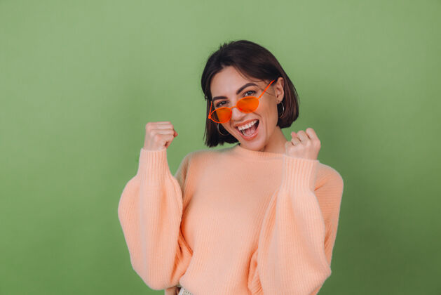 年轻年轻时尚的女士穿着休闲的桃色毛衣 戴着橙色眼镜 隔着绿橄榄色的墙壁积极地做着赢家的手势握紧拳头复制空间庆祝时尚赢家