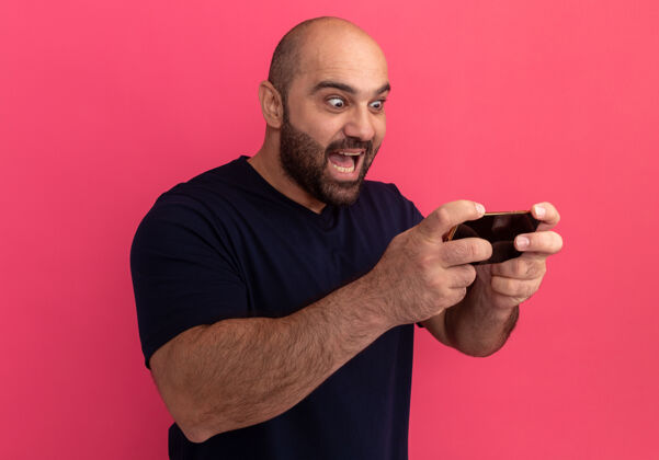 男人留着胡子的男人穿着海军蓝t恤 用智能手机玩游戏 站在粉色的墙上 看上去既惊讶又兴奋胡子站着兴奋