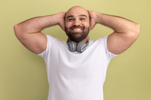 耳机一个留着胡子的男人 穿着白色t恤 戴着耳机 高兴地站在绿色的墙上 双手放在脑后胡子站着男人