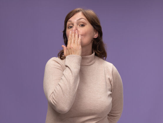 中年穿米色套头衫的中年妇女站在紫色的墙上吹吻吻女人站