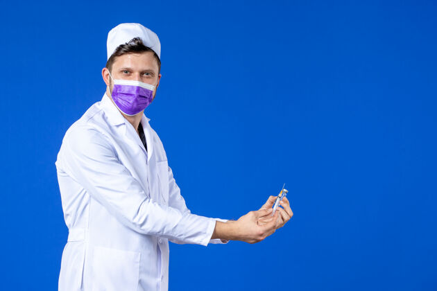 大流行前视图中的男医生穿着医疗服和口罩持有疫苗和注射在蓝色男医生药品注射