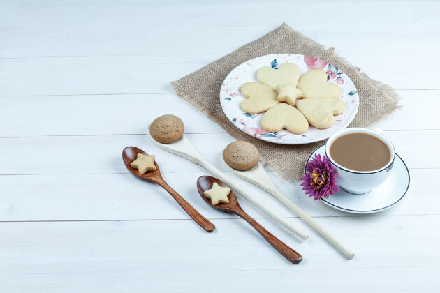烘焙高角度看心形饼干放在一个带花的袋子上 饼干放在木勺里 咖啡杯放在白色木板背景上水平形状早餐杯子