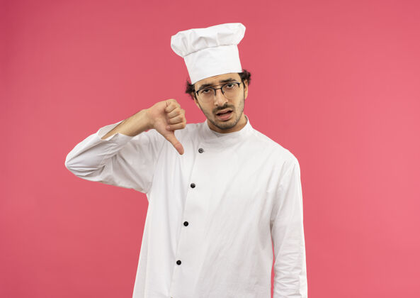 穿悲伤的年轻男厨师穿着厨师制服 戴着眼镜 拇指朝下粉色男人烹饪