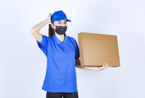 思想戴着面具 穿着蓝色制服的女快递员手里拿着一个大的纸板包裹 看上去困惑而犹豫不确定外卖服装