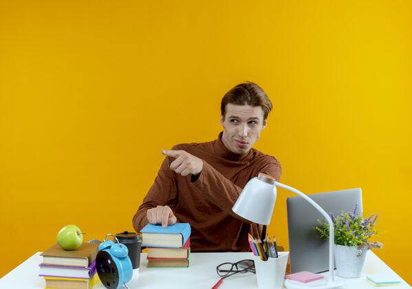 年轻看着边思考的年轻学生男孩坐在书桌旁 手里拿着学校的工具指着旁边点书桌黄色
