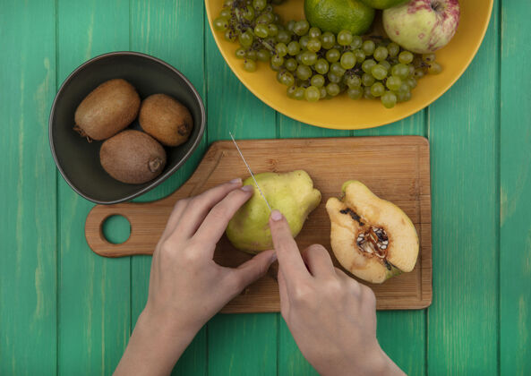 板俯瞰图：一个女人在砧板上切梨 在绿色的墙上切猕猴桃 葡萄和橘子梨女人食物