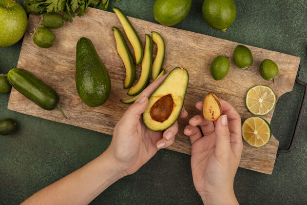 营养一只手拿着一个鳄梨 另一只手拿着它的果核的女性手的俯视图在一个木制的厨房板上 在绿色的表面上隔离着酸橙和绿色的苹果膳食手坑