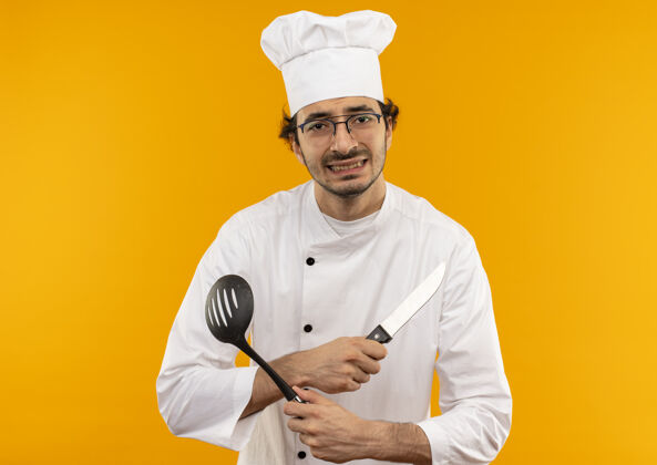 十字年轻的男厨师穿着厨师制服 戴着眼镜 双手交叉 手里拿着抹刀抹刀眼镜烹饪