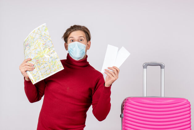 流行病戴着面具的年轻男子的正面图 手里拿着飞机票和白墙上的地图机票夏天面具