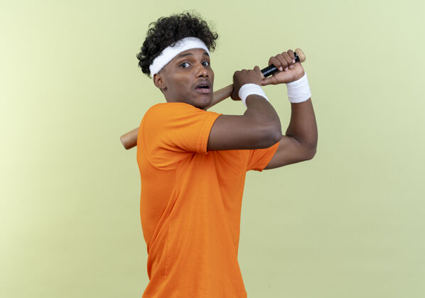 运动被吓坏了的年轻的美国黑人运动男子戴着头带和腕带把球棒放在肩膀上隔离在绿色的墙上腕带肩膀穿