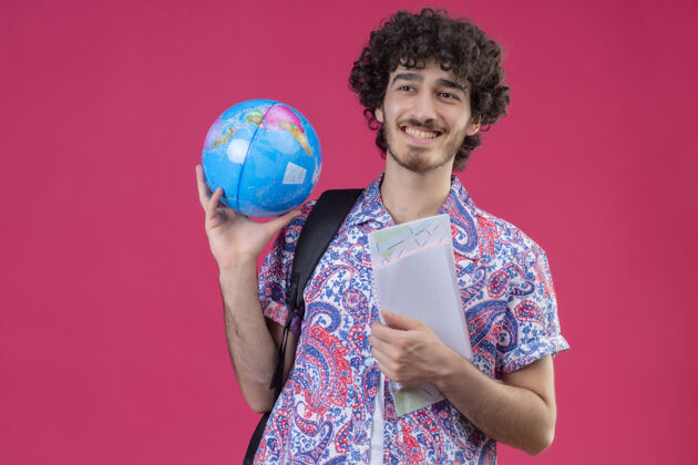 英俊微笑的年轻英俊的卷发旅行者男子拿着地球仪和笔记本在孤立的粉红色墙上与复制空间垫着地球仪年轻