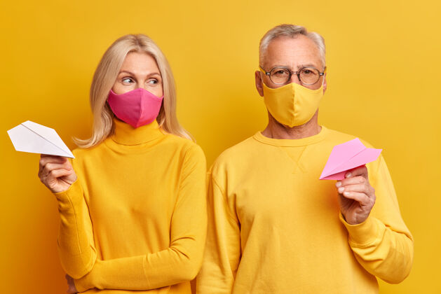 男人老年夫妇戴一次性口罩以防冠状病毒感染检疫期间待在家里穿着休闲黄色衣服手持手工纸飞机在工作室摆姿势飞机成熟病毒