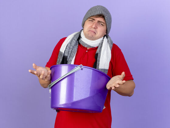帅哥悲伤的年轻英俊的金发病男子戴着冬天的帽子和围巾拿着塑料桶孤立在紫色的墙上男人手势悲伤