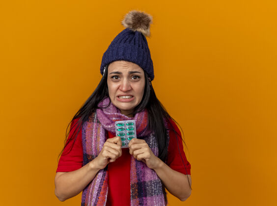 包恼怒的年轻白种人生病的女孩戴着冬天的帽子和围巾 手里拿着一包胶囊和帽子下的其他包 隔离在橙色的墙上 留着复印空间穿胶囊围巾