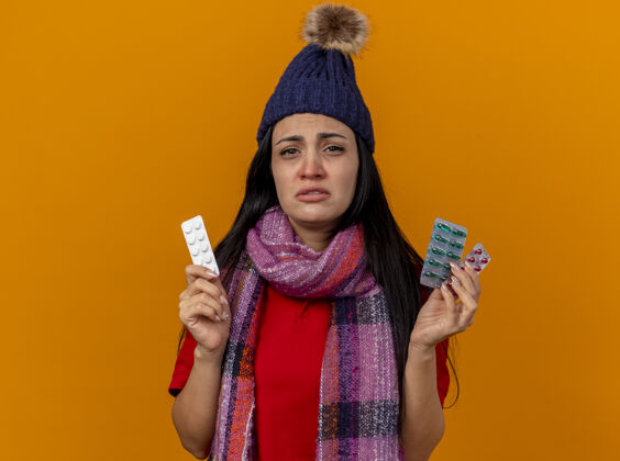 年轻虚弱的年轻白种病女孩戴着冬天的帽子 戴着围巾 手里拿着几包药片 隔离在橙色的墙上 留着复印空间穿虚弱橙色