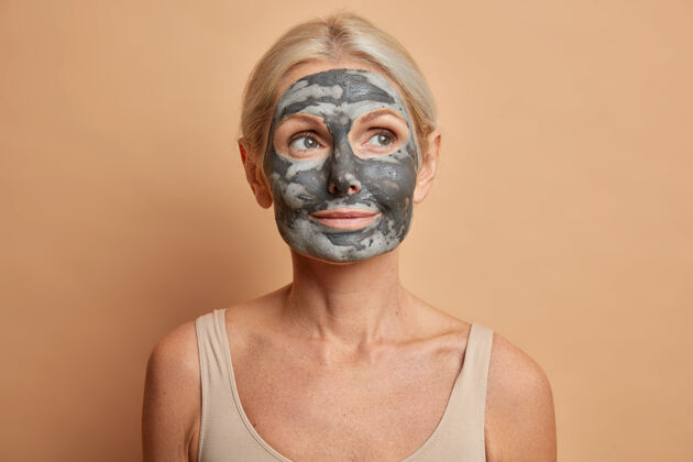 治疗贴心的欧洲女人的特写镜头涂上泥面膜 把目光移开关心肤色的姿势 光着肩膀隔离在棕色的墙上自然老年提举