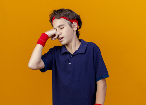 头带疼痛的年轻英俊的运动男孩戴着头带和带牙套的腕带把手放在眼睛上闭着眼睛隔离在橙色的墙壁上 有复制空间男孩橙色腕带