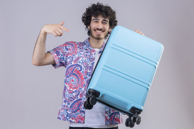 微笑微笑着的年轻英俊的卷发旅行者 拿着手提箱 指着孤零零的白墙上的手提箱年轻男人抱着