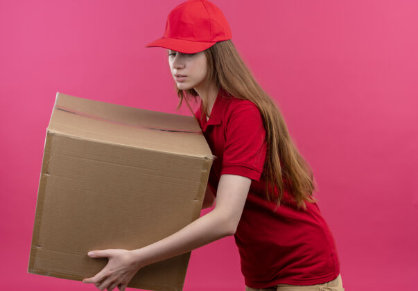 女人穿着红色制服的年轻送货女孩拿着箱子 俯视着 站在孤立的粉红色墙上的侧视图里制服按住视野