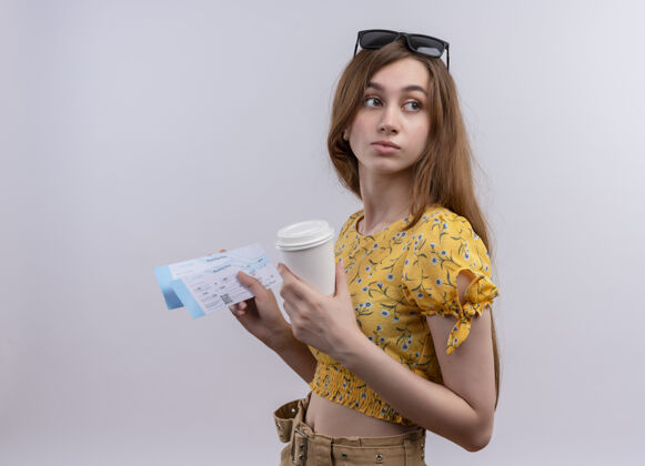 穿年轻的旅行女孩头戴太阳镜 拿着飞机票和塑料咖啡杯 站在隔离的白色墙壁上看侧面图中的票太阳镜轮廓女孩