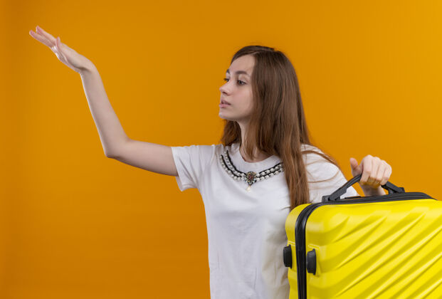举起年轻的旅行家女孩拿着手提箱 在孤立的橙色墙上举起左手年轻人左边拿着