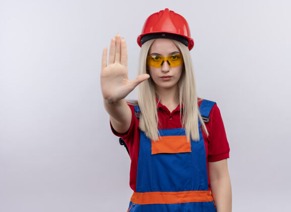 手势年轻的金发工程师 穿着制服 戴着安全眼镜的建筑工女孩 伸出手 在隔离的白色墙壁上做手势 留有复印空间安全金发制服