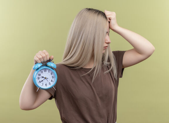 压力紧张的金发女孩拿着闹钟把手放在孤立的绿色墙上抱着手女孩