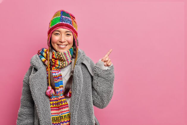 辫子快乐的亚洲女人冬季服装寒冷的天气表明在空白处显示一些可怕的穿针织帽子皮衣和围巾围脖隔离在粉红色的墙壁女性广告漂亮