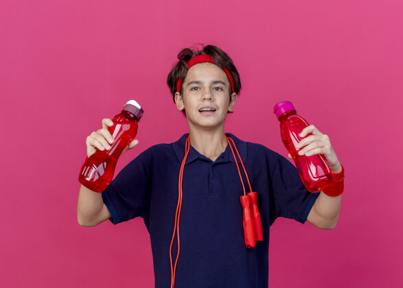 年轻印象深刻的年轻英俊的运动男孩戴着头带和护腕与牙套和跳绳脖子上举行的深红色墙上孤立的水瓶腕带周围背带