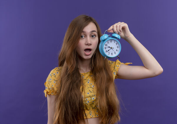 女孩惊讶的年轻女孩拿着闹钟在孤立的紫色墙上惊喜抱着年轻