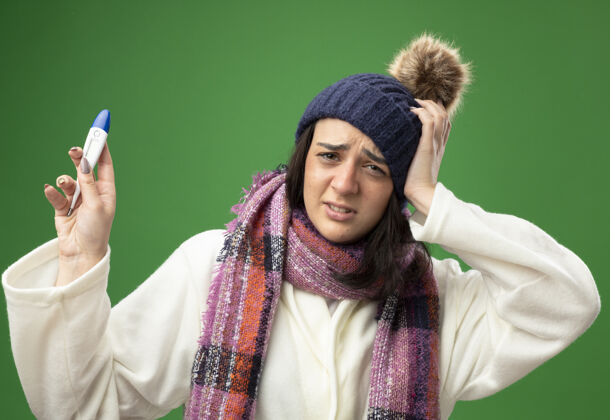 头疼痛的年轻白种人生病的女孩穿着长袍 戴着冬天的帽子和围巾 手里拿着温度计 手放在头上 隔离在绿色的墙上穿冬天长袍