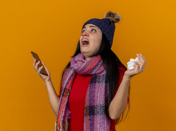 手机年轻的高加索病女孩戴着冬天的帽子和围巾 拿着手机和餐巾 孤零零地看着橙色的墙上年轻帽子不愉快