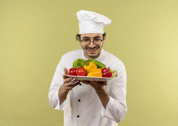 男性面带微笑的年轻男厨师穿着厨师制服 戴着眼镜 拿着蔬菜看着隔离在绿墙上的盘子微笑蔬菜盘子
