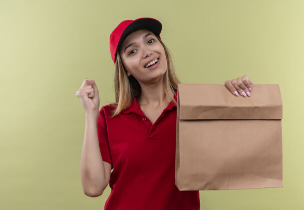 微笑微笑着的年轻送货女孩穿着红色制服 戴着帽子 手里拿着纸袋 在绿色的墙壁上展示着“是”的手势帽子包抱着