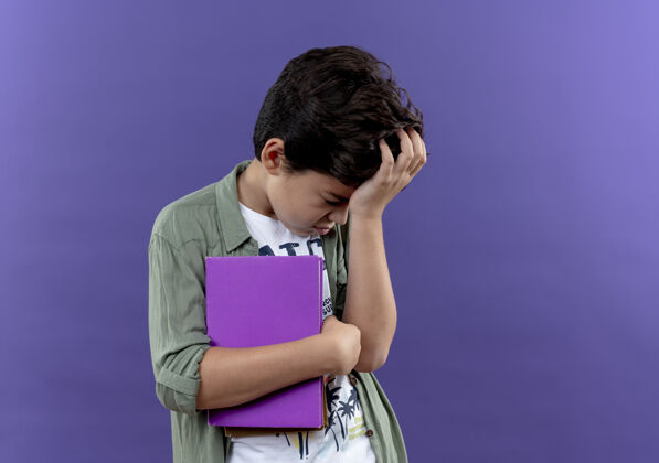 小低着头悲伤的小男生拿着书 把手放在头上 隔离在紫色的墙上 留着复印空间头小学生抱
