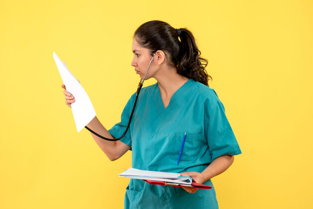 肖像穿着制服的女医生正看着黄色墙上夹着文件夹的文件文件夹快乐自信