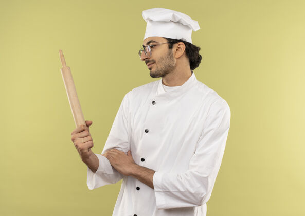 男高兴的年轻男厨师穿着厨师制服 戴着眼镜 拿着擀面杖看着隔离在绿色墙上的擀面杖拿着滚年轻