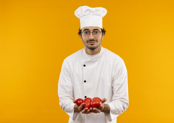 手持年轻的男厨师穿着厨师制服 戴着眼镜 手里拿着隔离在黄色墙上的西红柿年轻男性制服
