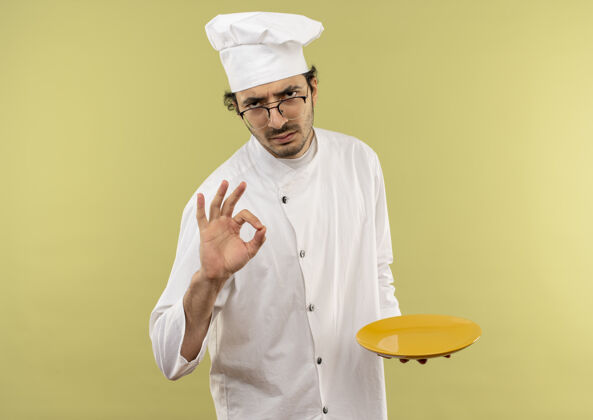 严格严格的年轻男厨师穿着厨师制服 戴着眼镜拿着盘子 在绿色的墙壁上显示出奥基的手势盘子拿着穿
