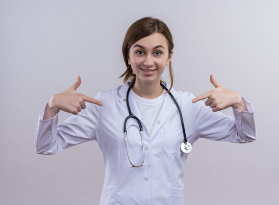 长袍年轻的女医生面带微笑 穿着医用长袍 戴着听诊器 指着孤立的白墙上的自己女人听诊器指点
