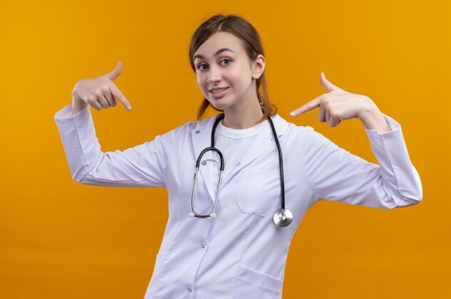 长袍面带微笑的年轻女医生穿着医用长袍 手持听诊器 指着孤立的橙色墙上的自己听诊器指点医生