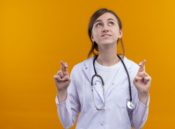 医生希望年轻的女医生穿着医用长袍和听诊器 用交叉的手指看着隔离的橙色墙壁上的复制空间女女人愿望