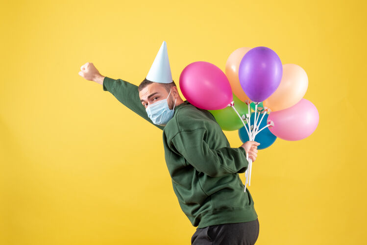 生日正面图：黄色背景上戴着面具手持彩色气球的年轻男性大流行气球聚会