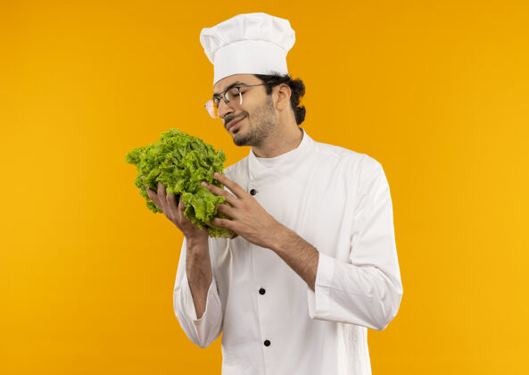 持有年轻的男厨师闭着眼睛 穿着厨师制服 戴着眼镜 手里拿着隔离在黄色墙上的沙拉请佩戴厨师