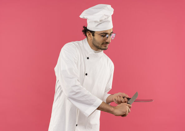 年轻高兴的年轻男厨师穿着厨师制服和眼镜磨刀与切肉刀隔离在粉红色的墙上男性烹饪刀