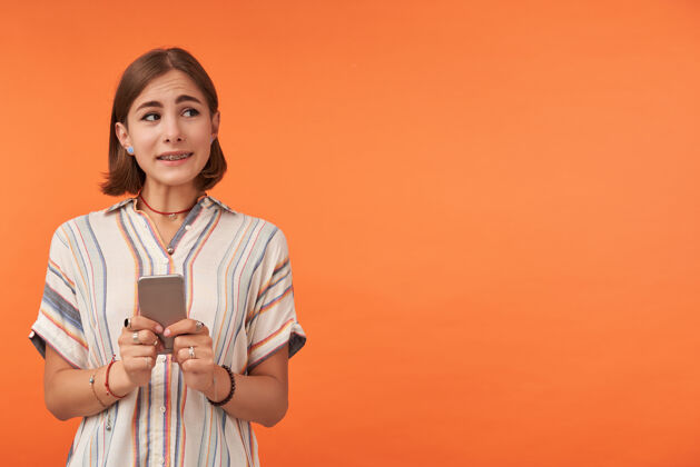 休闲年轻可爱的女孩拿着一部智能手机 看着右边橙色墙上的复制空间 看起来很尴尬穿着条纹衬衫 背带 项链和手镯哦女士社交