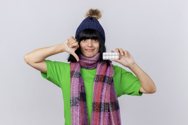 相机戴着冬日帽子和围巾的年轻白种人女孩看着相机 镜头里是一包平板电脑 拇指朝下 背景是白色的 有复印空间药片下来年轻