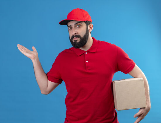 帅气自信的年轻英俊的白人送货员穿着红色制服 戴着帽子 手里拿着纸箱 手上空空如也的蓝色隔离空自信交货