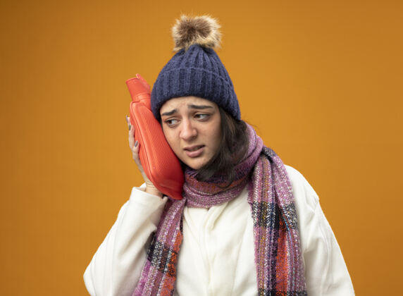 壁板虚弱的年轻白种人生病的女孩穿着长袍 戴着冬天的帽子和围巾 用热水袋抚摸着头 看着橙色背景上孤立的一面 还有复制空间女孩围巾热