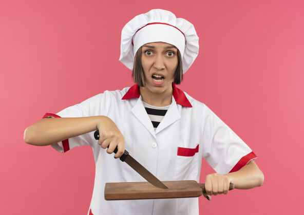 板令人印象深刻的年轻女厨师在厨师制服触摸切菜板与刀隔离在粉红色年轻触摸女